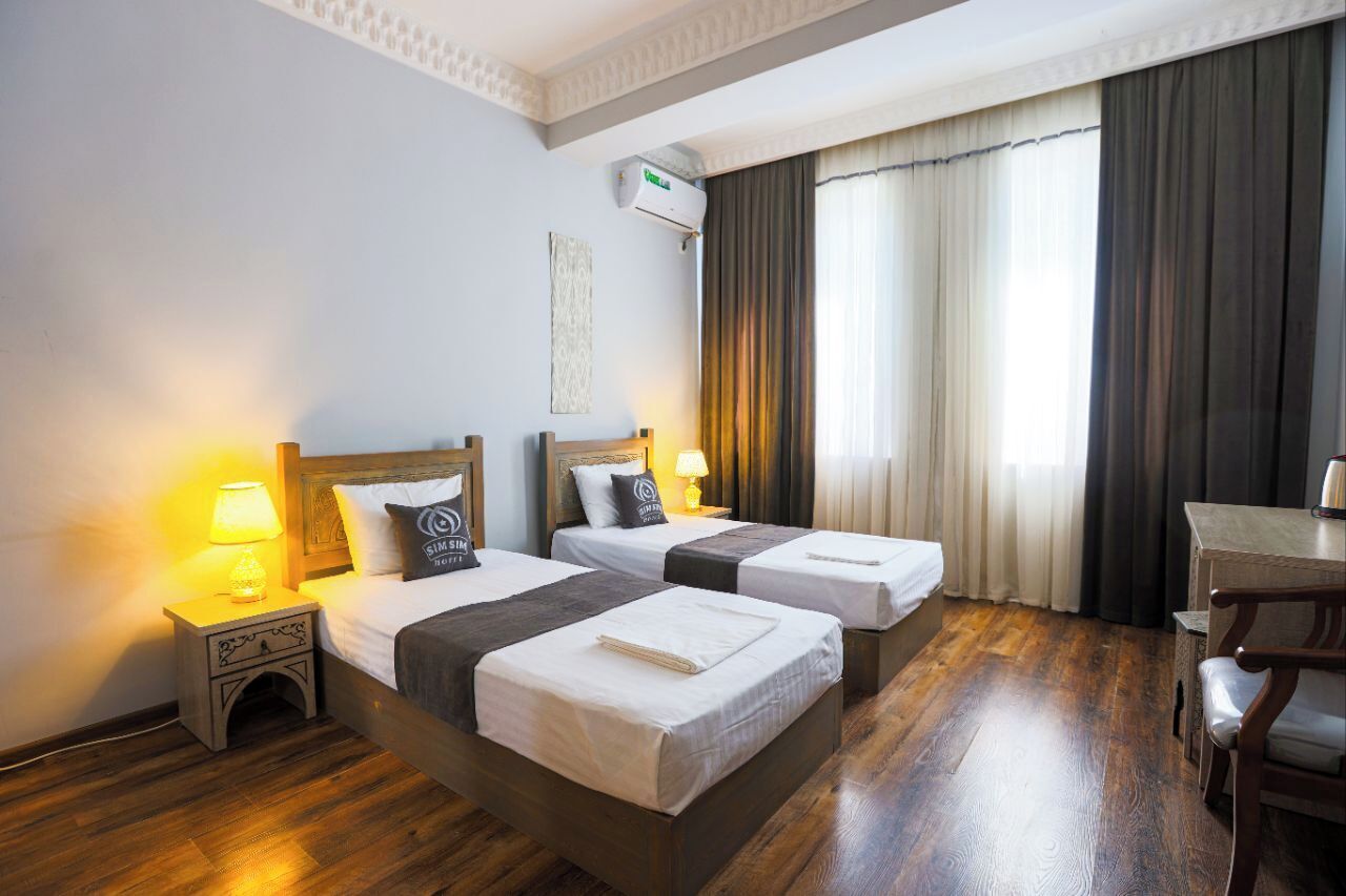 A hotel room with two beds, a desk, a chair, an air conditioning unit, and a window with curtains.