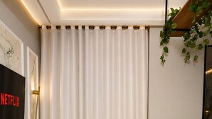Uma sala de estar moderna com um sofá bege, televisão na parede e uma grande janela com cortinas.