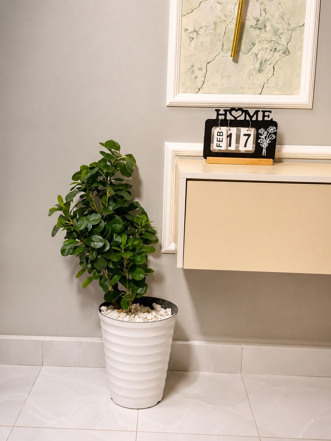 Ein Blumentopf mit weißen Kieselsteinen, eine gerahmte Karte an der Wand und ein schwarzer Kalender mit 'HOME' und dem Datum '17' auf einem beigefarbenen Schrank.