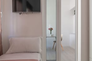 Ein Schlafzimmer mit einem Bett, einem an der Wand befestigten Fernseher und Blick in einen weiteren Raum mit Tisch und Stuhl.