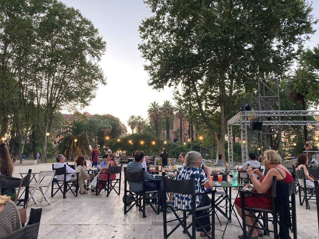 An outdoor seating area with tables and chairs, surrounded by trees and a stage structure.