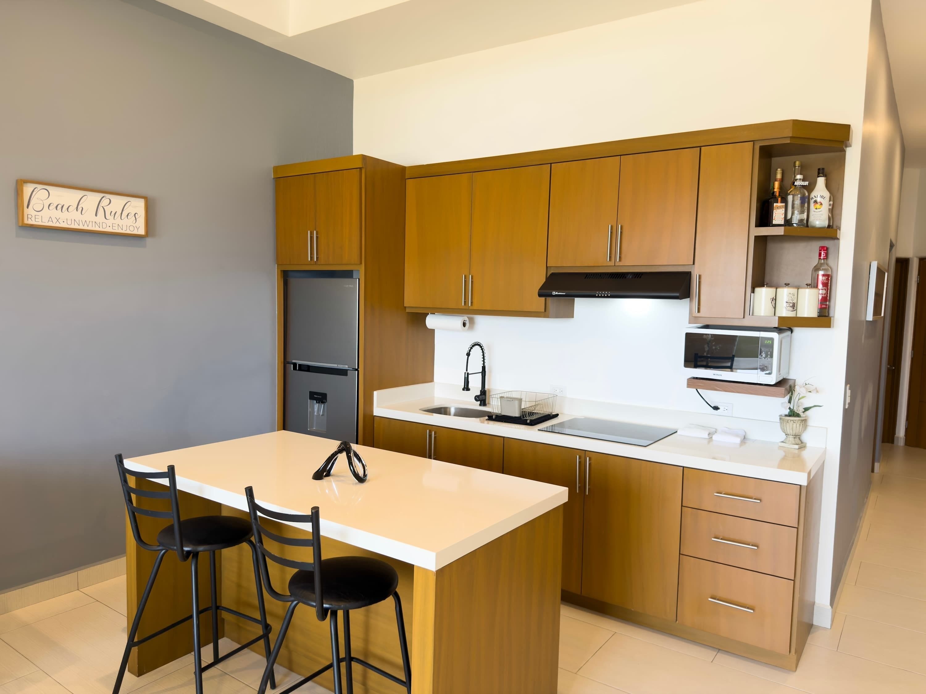 A modern kitchen with wooden cabinets, a stainless steel refrigerator, a microwave, and a sink. There are two bar stools and a small island with a sink.