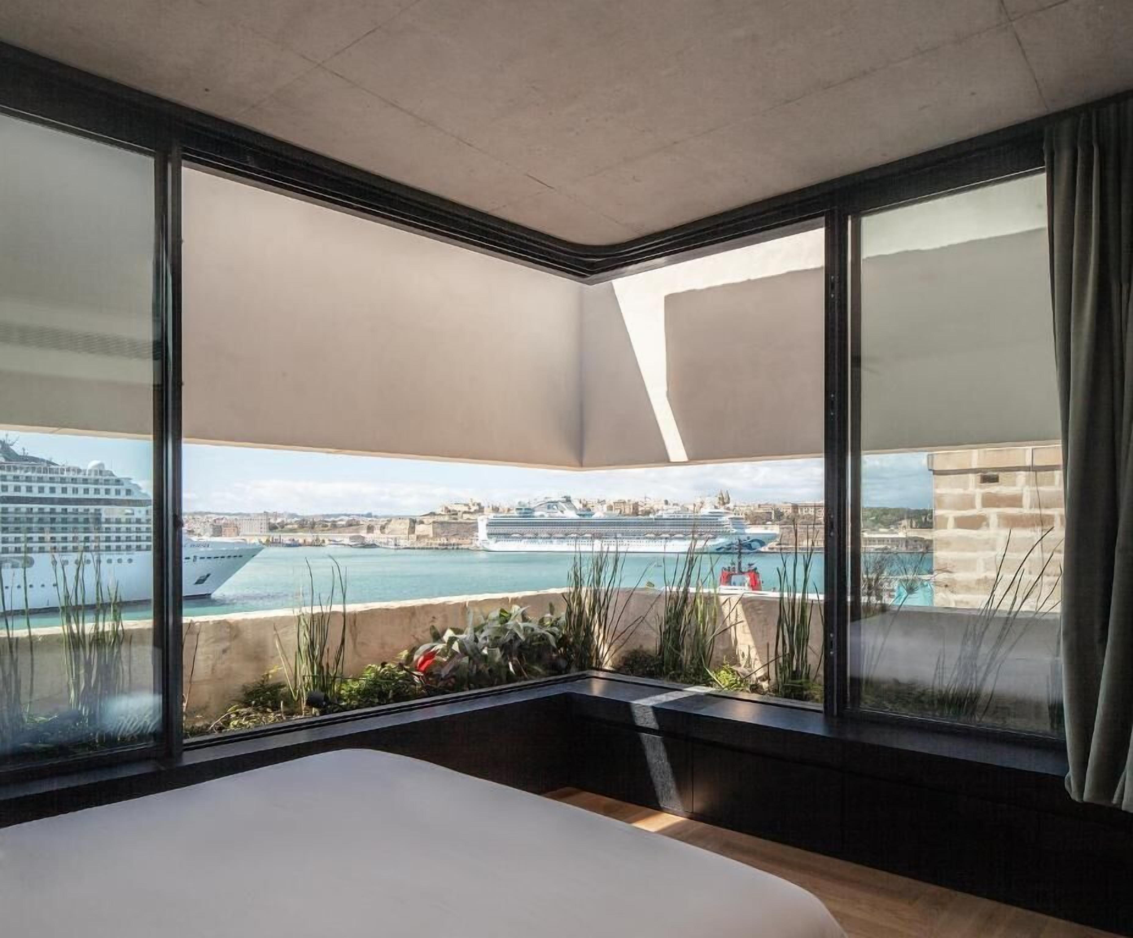 Habitación de hotel con una cama grande, vista a un puerto con cruceros y un balcón con plantas.