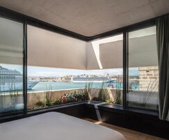 Quarto de hotel com uma cama grande, vista para um porto com navios de cruzeiro e uma varanda com plantas.
