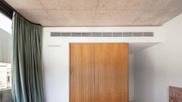 A bedroom with a wooden wardrobe, a bed, a window with curtains, and a view of a building outside.