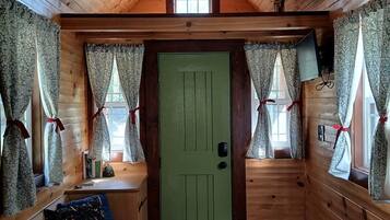 Ein Interieur einer Holzhütte mit grüner Tür, Holzwänden, einem Bett, einem Schreibtisch und einem Fenster mit Vorhängen.