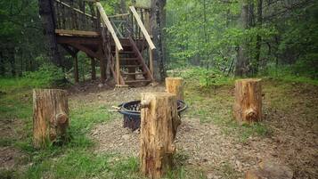 Une terrasse en bois rustique, dotée d’un escalier menant à une plateforme, est entourée d’une zone boisée parsemée de plusieurs souches d’arbres.