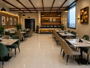 A restaurant with a long dining area, multiple tables, and chairs. There is a counter with a display of food items and a view of the city through large windows. - Hotel Ocean Pearl (Agra)
