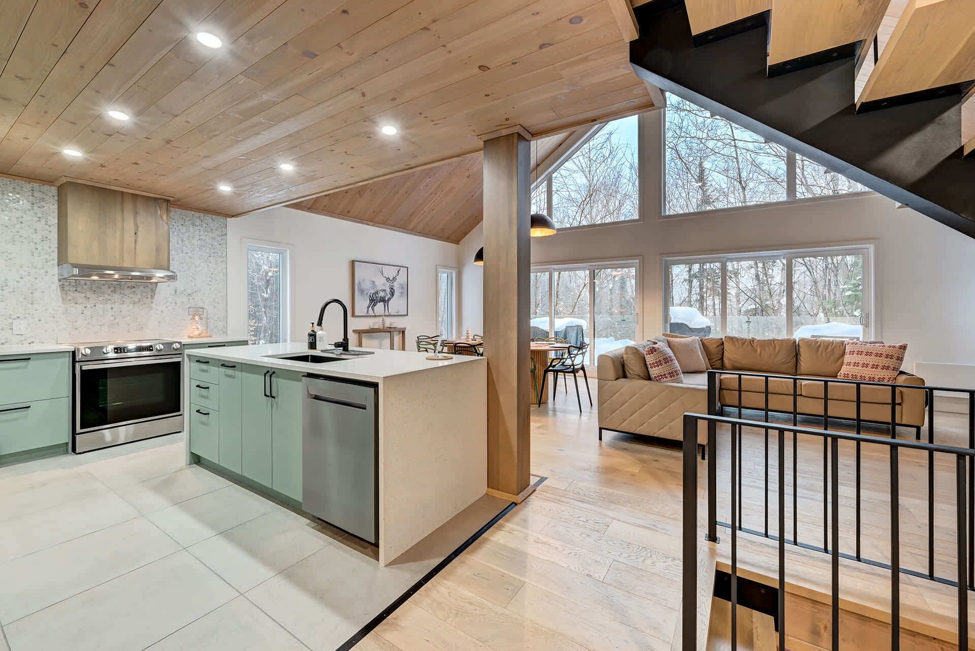 A modern kitchen with a central island, a dining area with a sofa, and large windows offering views of the outdoors.