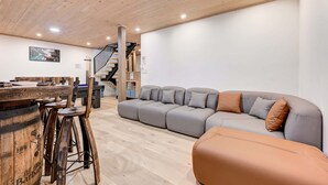 Ein modernes Wohnzimmer mit Holzbalkendecke, einem grauen Ecksofa, einer Holzbar mit Barhocker und einer Treppe.