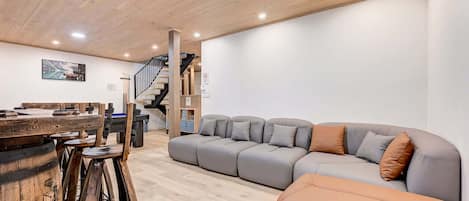 A modern living room with a wooden ceiling, a grey sectional sofa, a wooden bar with stools, and a staircase.