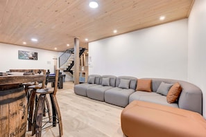 A modern living room with a wooden ceiling, a grey sectional sofa, a wooden bar with stools, and a staircase. - Nayati Chalet | Luxury Chalet with Spa Near Mont-Tremblant (Amherst)