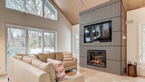 Ein modernes Wohnzimmer mit einem großen Sofa, einem Kamin, einem Flachbildfernseher und großen Fenstern.