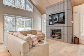 A modern living room with a large sofa, a fireplace, a flat-screen TV, and large windows. - Nayati Chalet | Luxury Chalet with Spa Near Mont-Tremblant (Amherst)