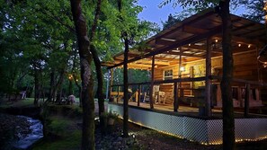 Una cabaña de madera con terraza, rodeada de árboles y un arroyo.
