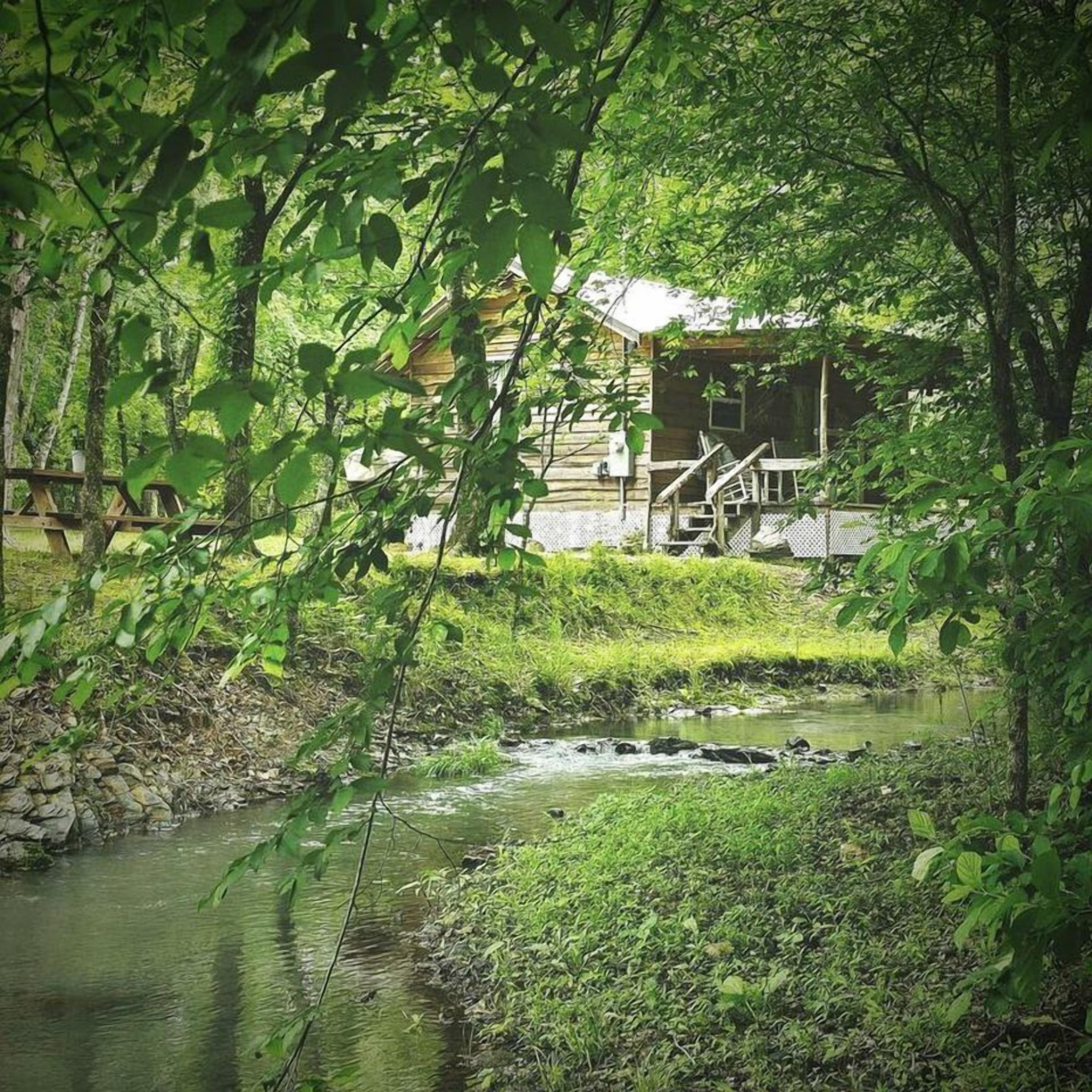 A rustic cabin with a wooden deck and a small stream flowing in front.