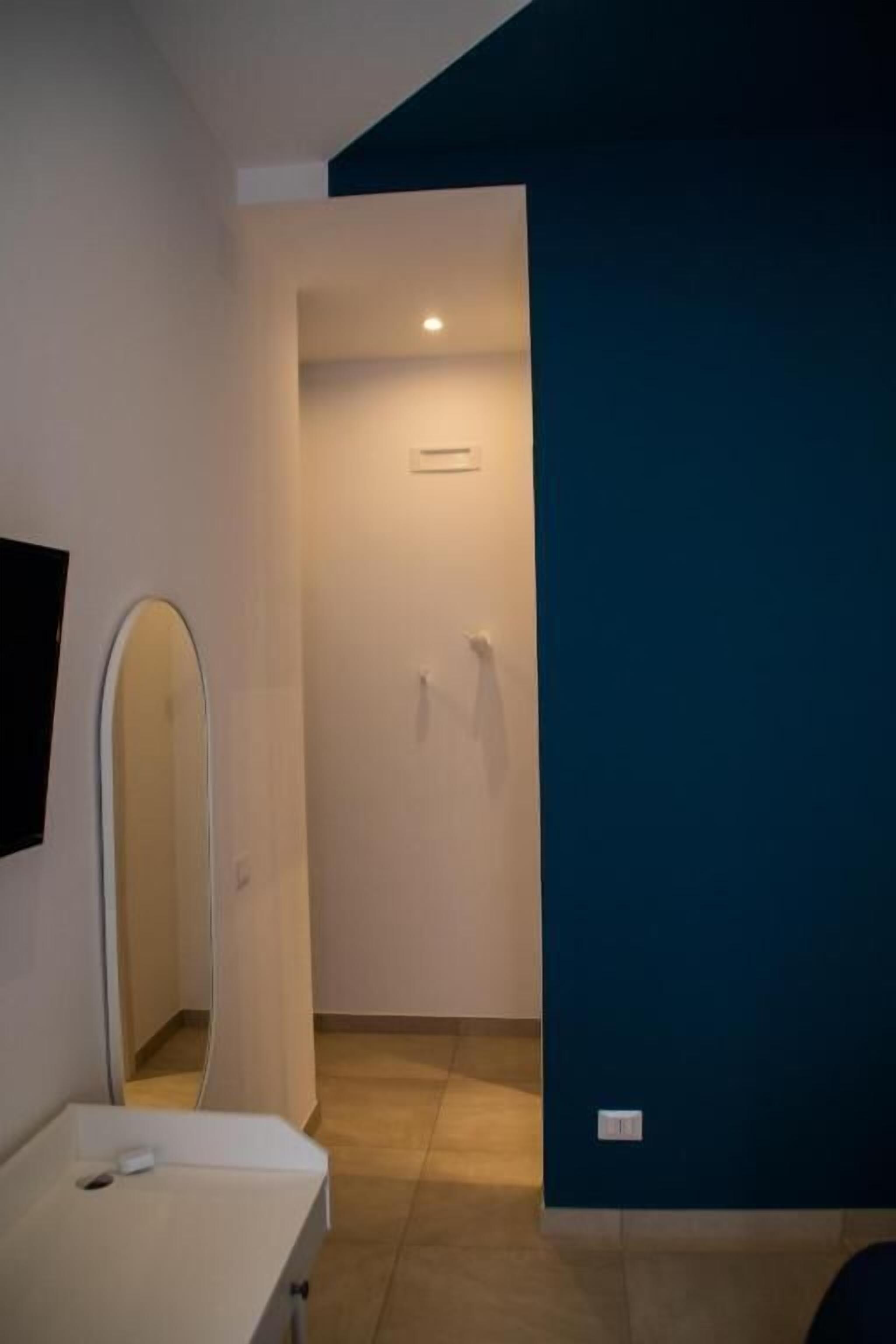 A hotel room with a white bedside table, a mirror, a door leading to another room, and a blue accent wall.