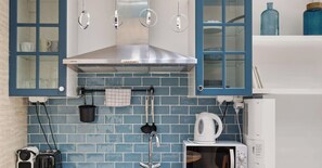 A kitchen with blue tiles, a stainless steel range hood, hanging kitchen utensils, and a microwave. - Elégant 2 pièces proche du Marais (Paris)