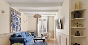 Ein Wohnzimmer mit einem blauen Sofa, einem kleinen Tisch und einem Sessel. An der Wand hängt ein großes Bild in einem Rahmen und ein Fernsehgerät ist an der Wand montiert.