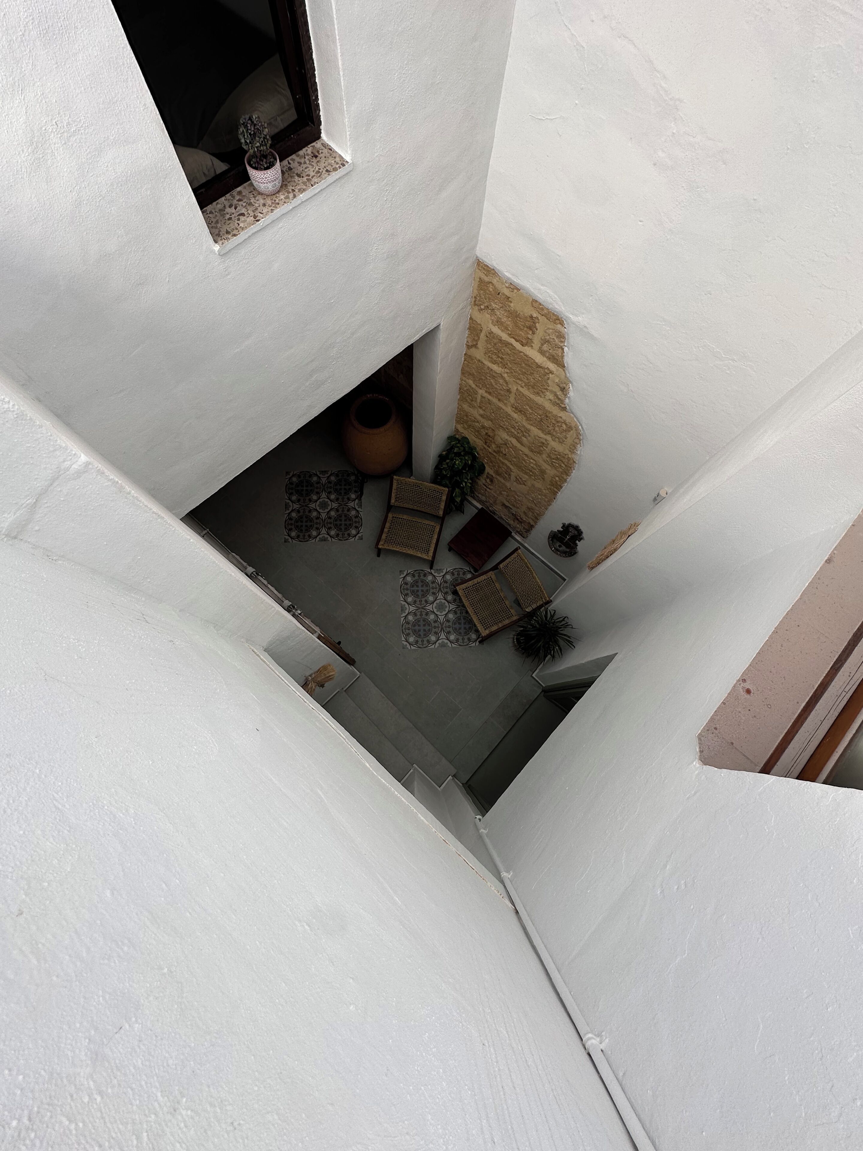 Una escalera estrecha con paredes blancas y una pared de piedra a un lado. Hay plantas en macetas, una cesta tejida y un piso de baldosas.