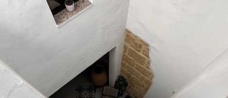 Una escalera estrecha con paredes blancas y una pared de piedra a un lado. Hay plantas en macetas, una cesta tejida y un piso de baldosas.