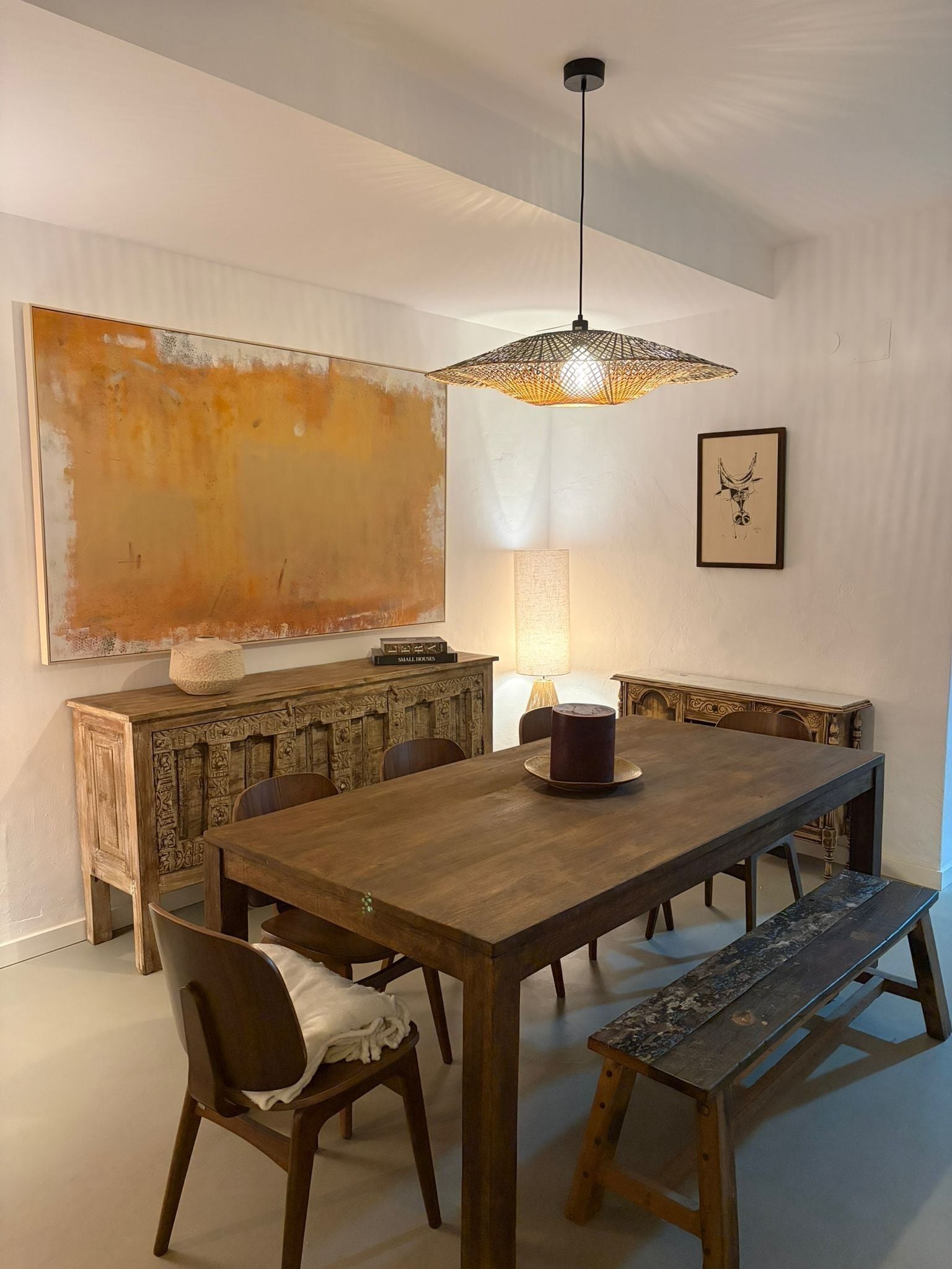 A dining area with a wooden table, benches, chairs, a hanging light fixture, a wall-mounted lamp, and a large abstract painting.