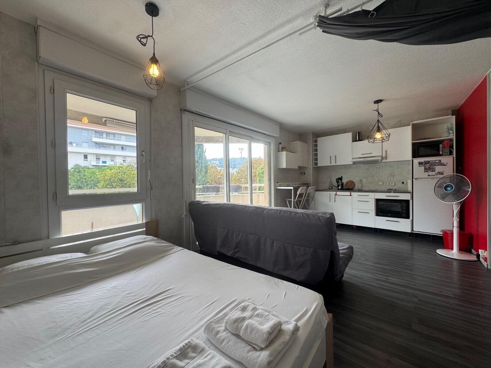 A bedroom with a bed, a window, and a kitchenette in the background.