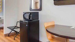 Habitación compacta con televisor de pantalla plana en la pared, un refrigerador negro pequeño, una mesa de madera con una silla, y una silla plegable de metal.