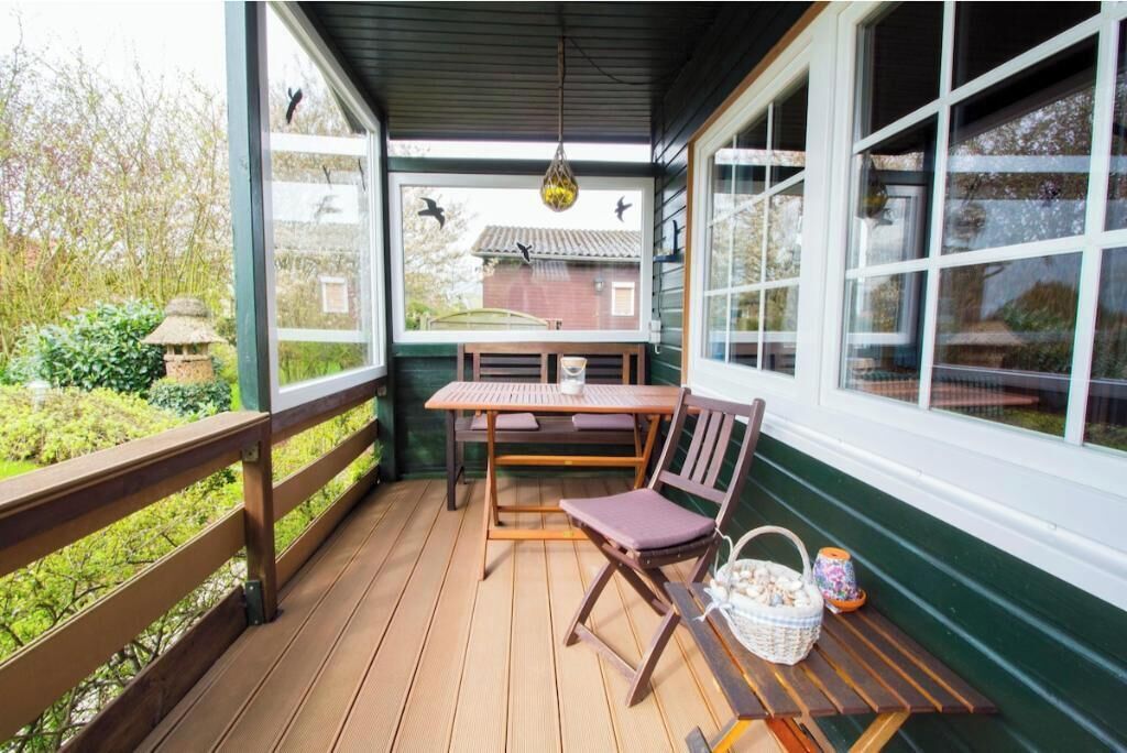 Een overdekt terras met een tafel, stoelen, een hanglamp en uitzicht op een huis door de ramen.