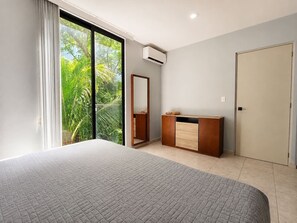 Ein Schlafzimmer mit einem großen Bett, einem Kleiderschrank, einem Spiegel und einer Glastür zum Balkon mit Grünpflanzen.