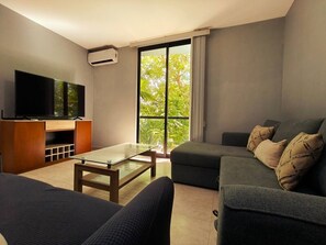 A modern living room with a flat-screen TV, a glass-top coffee table, a sofa, and a large window with a view of greenery. - Quiet 2BR Haven with Worspace Surrounded by Nature (Playa del Carmen)