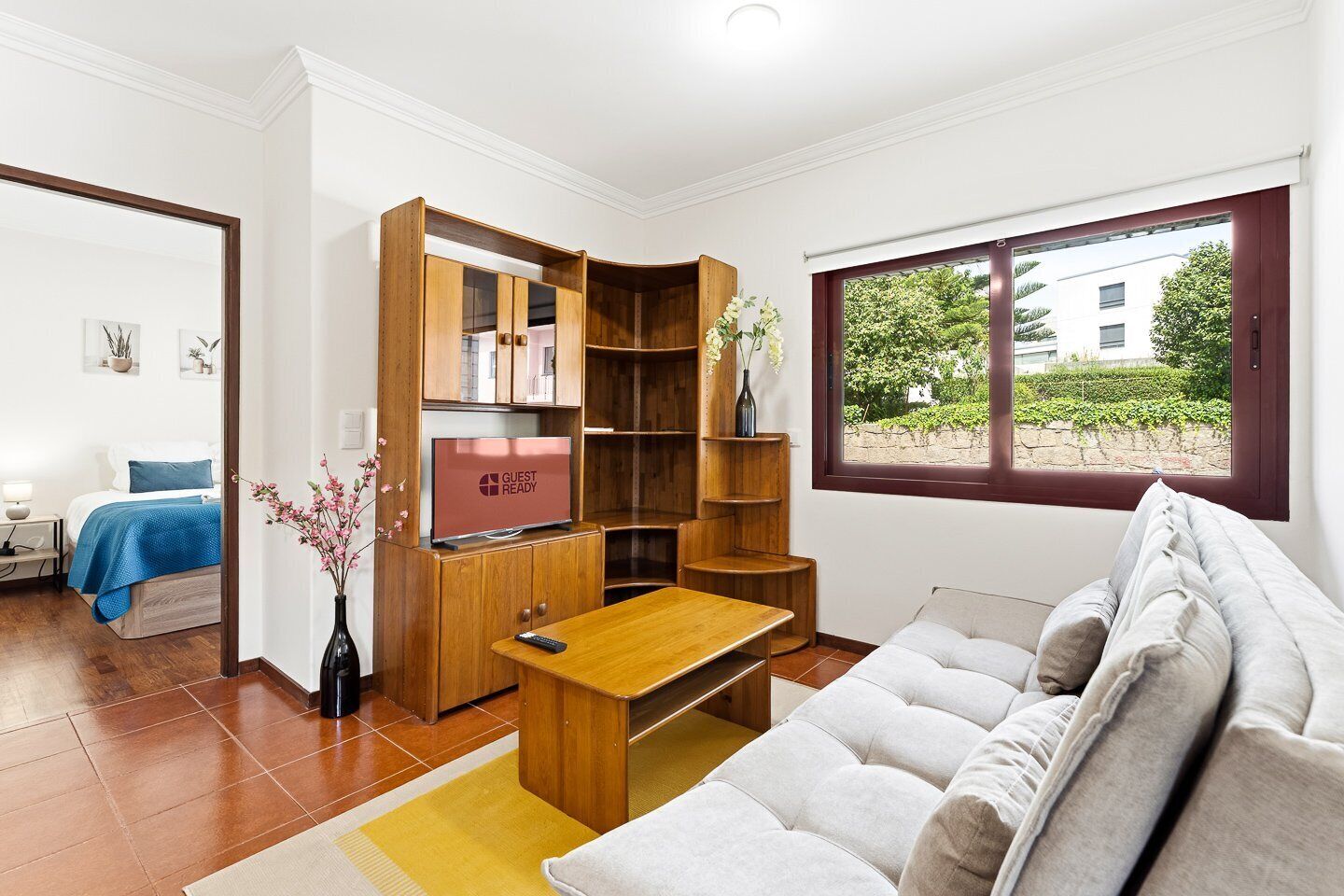 Ein modernes Wohnzimmer mit einer Couch, einem Holz-TV-Schrank, einem Esstisch und einem großen Fenster mit Blick auf Grünflächen und Gebäude.