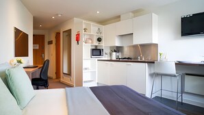 Ett modernt hotellrum med en säng, ett skrivbord, en stol, ett litet kök och en TV.