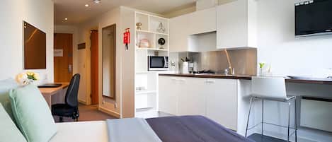 A modern hotel room with a bed, a desk, a chair, a kitchenette, and a TV.