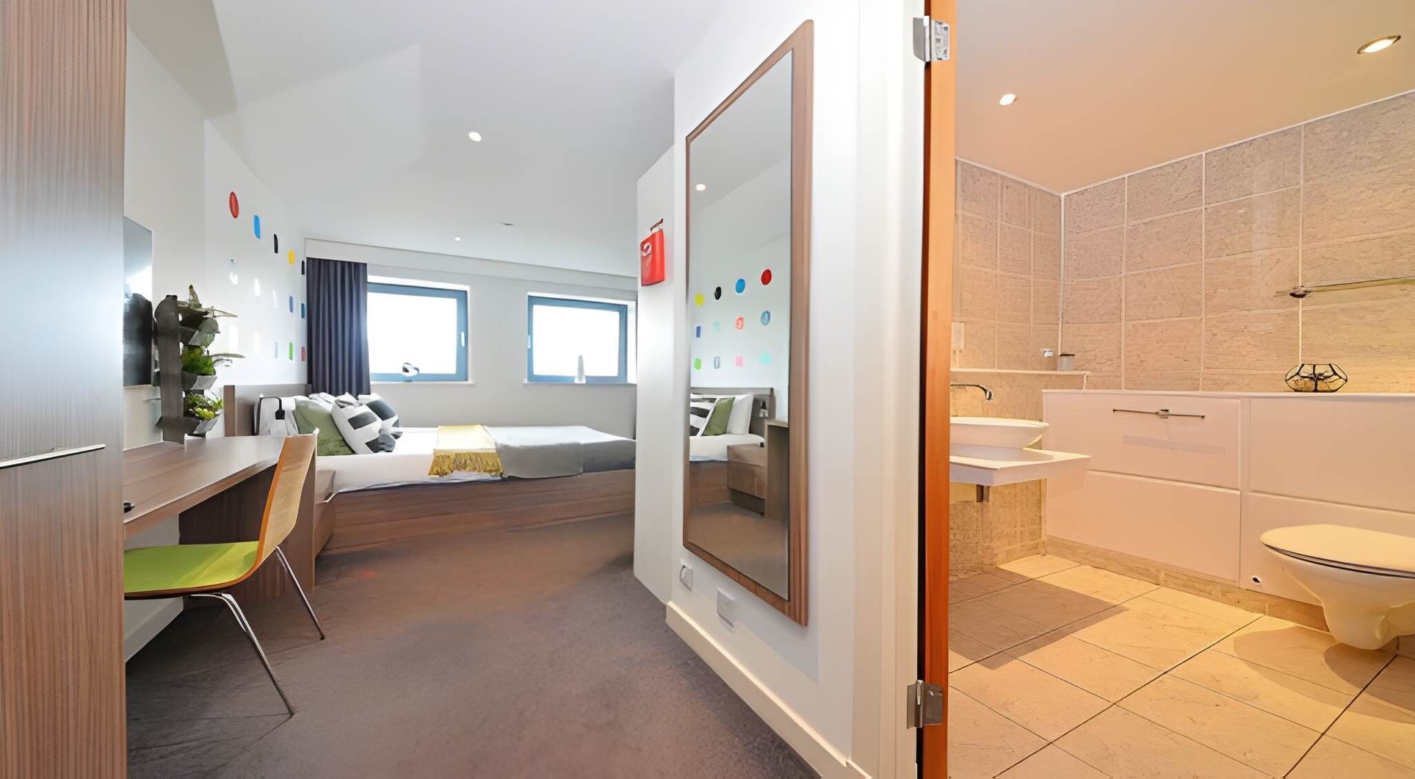 Una habitación de hotel moderna con cama, escritorio, silla, un espejo grande y un baño con inodoro y lavabo.