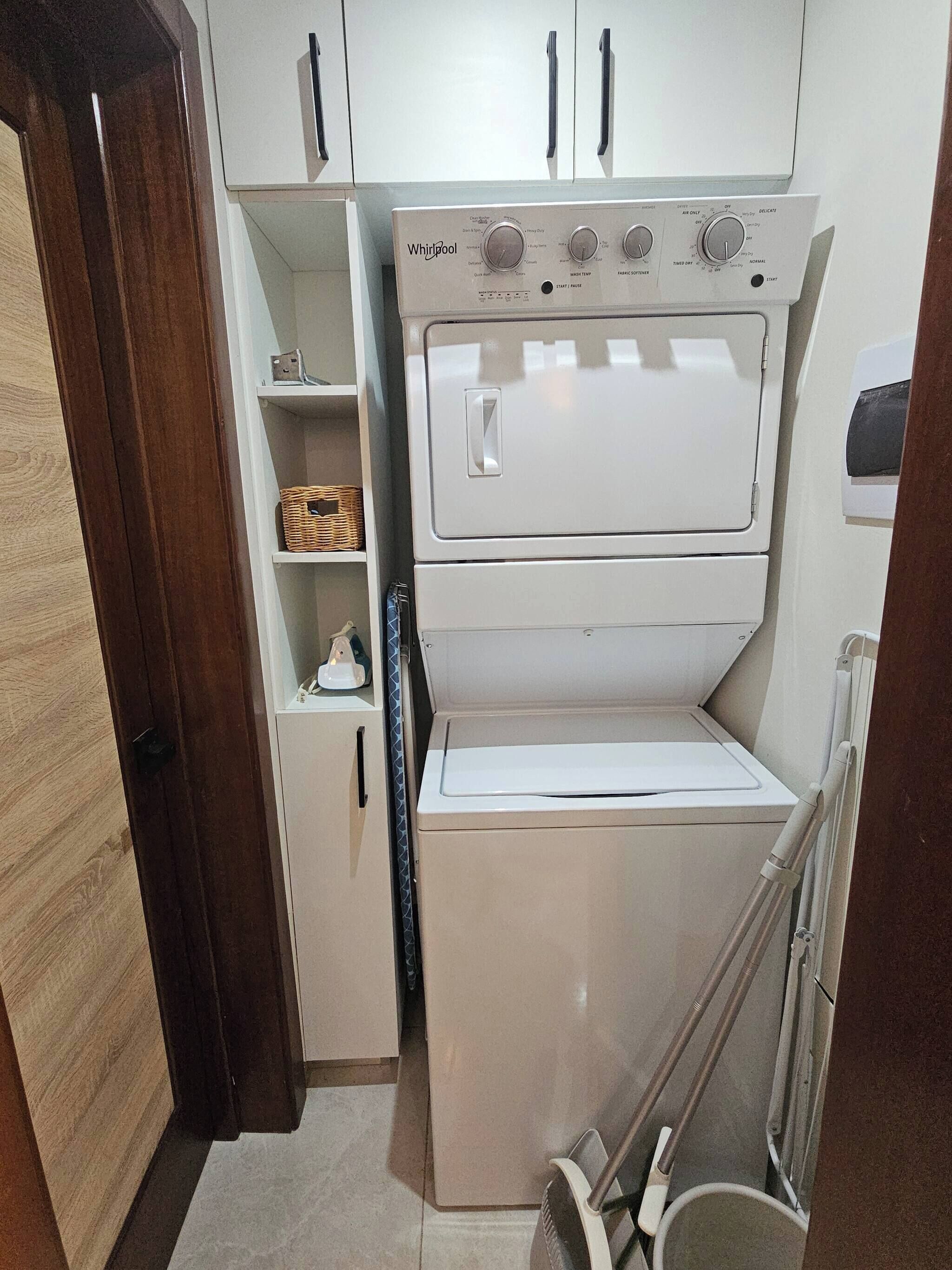 A compact laundry room with a Whirlpool washer and dryer, wooden cabinets, and a small shelf with a basket.