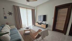 A living room with a sofa, a coffee table, a TV, and a door. - Eden Tropical new 1BR Condo, Walk to Beach ,pool (Punta Cana)