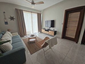 A living room with a sofa, a coffee table, a TV, and a door. - Eden Tropical new 1BR Condo, Walk to Beach ,pool (Punta Cana)