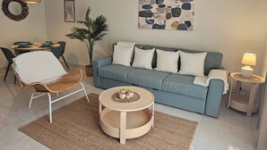 A living room with a blue sofa, a round coffee table, a wicker chair, a dining table with blue chairs, and a wall art piece. - Eden Tropical new 1BR Condo, Walk to Beach ,pool (Punta Cana)