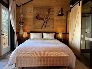 A large bed with a headboard, two bedside lamps, a woven bench, and a wooden wall with a bird painting. - Two Pine Cabin (Rocky View County)