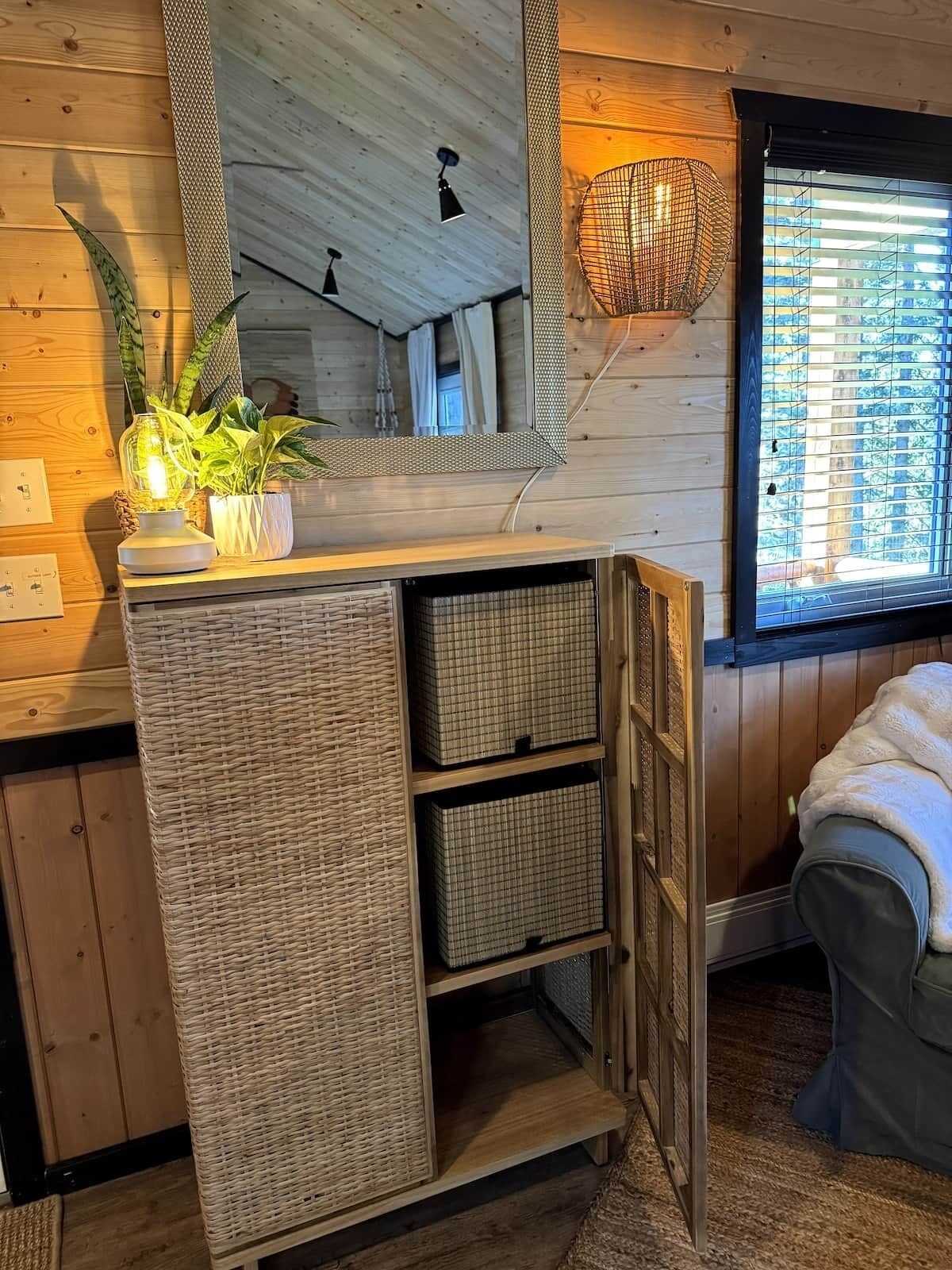 A wooden interior with a mirrored cabinet, wicker baskets, a potted plant, and a window with blinds.