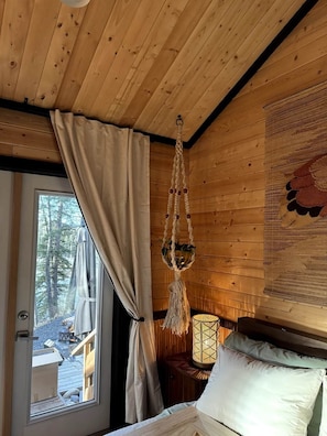 A wooden cabin interior with a bed, a macrame hanging light, and a view of the outdoors through a door. - Two Pine Cabin (Rocky View County)