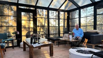A covered outdoor seating area with a fire pit, wooden furniture, and large windows offering a view of trees.