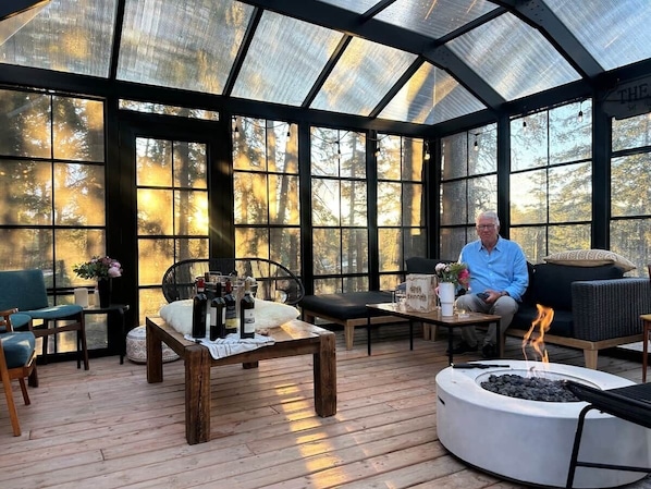 A covered outdoor seating area with a fire pit, wooden furniture, and large windows offering a view of trees.
