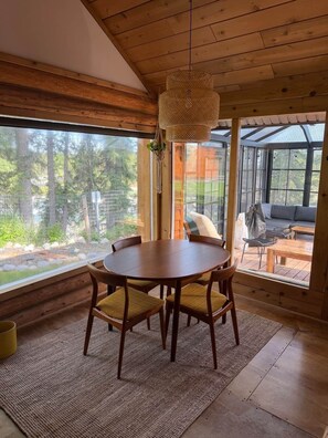 A wooden interior with a round table and chairs, large windows, and a hanging light fixture.