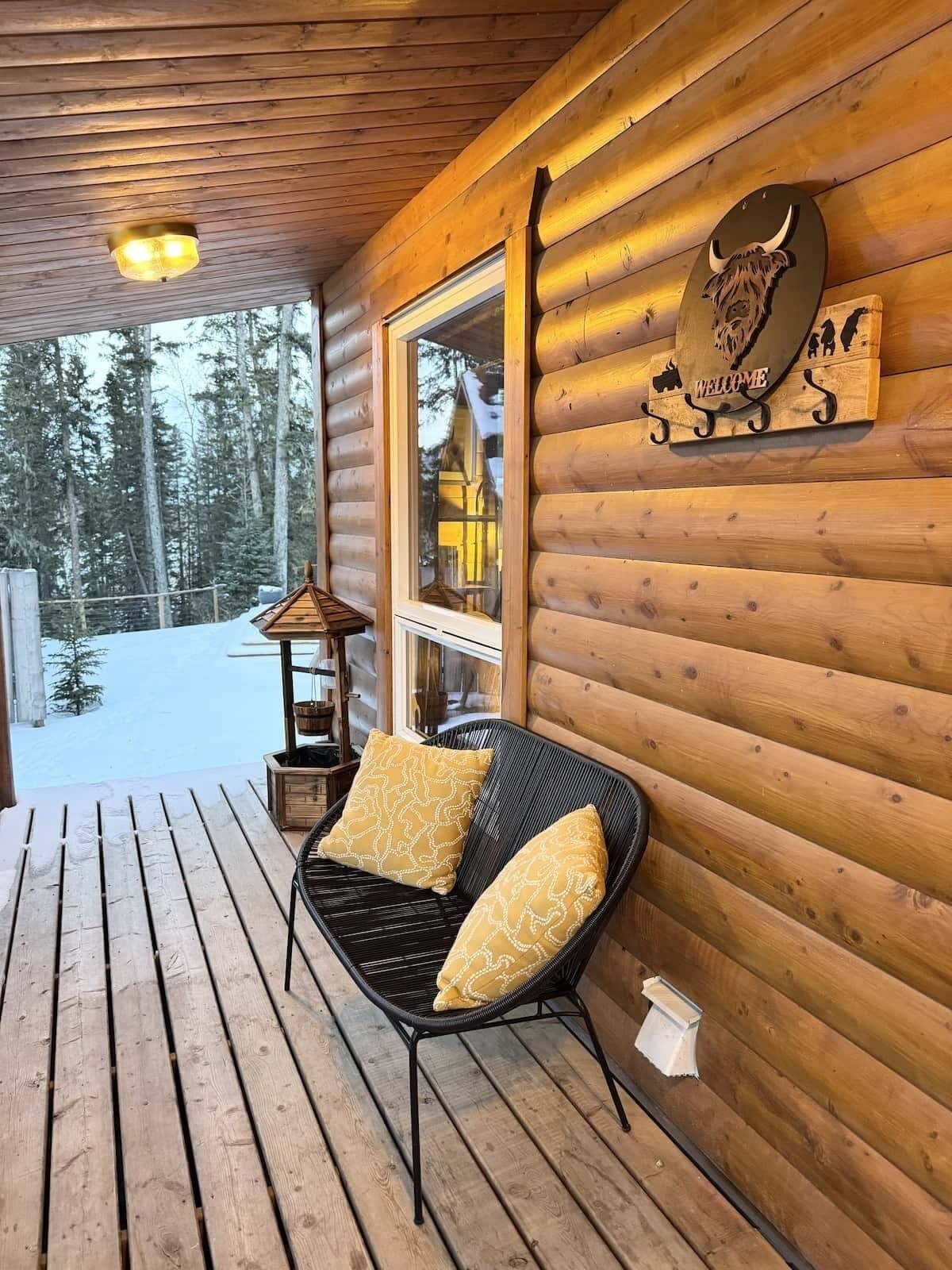 A wooden cabin with a porch, a black wicker chair with cushions, a welcome sign, and a view of a snowy forest.