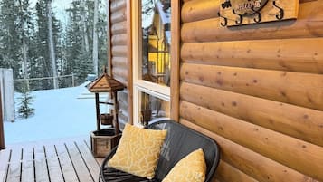 En stuga av trä med en veranda, en svart flätstol med kuddar, en välkomnande skylt och utsikt över en snötäckt skog.