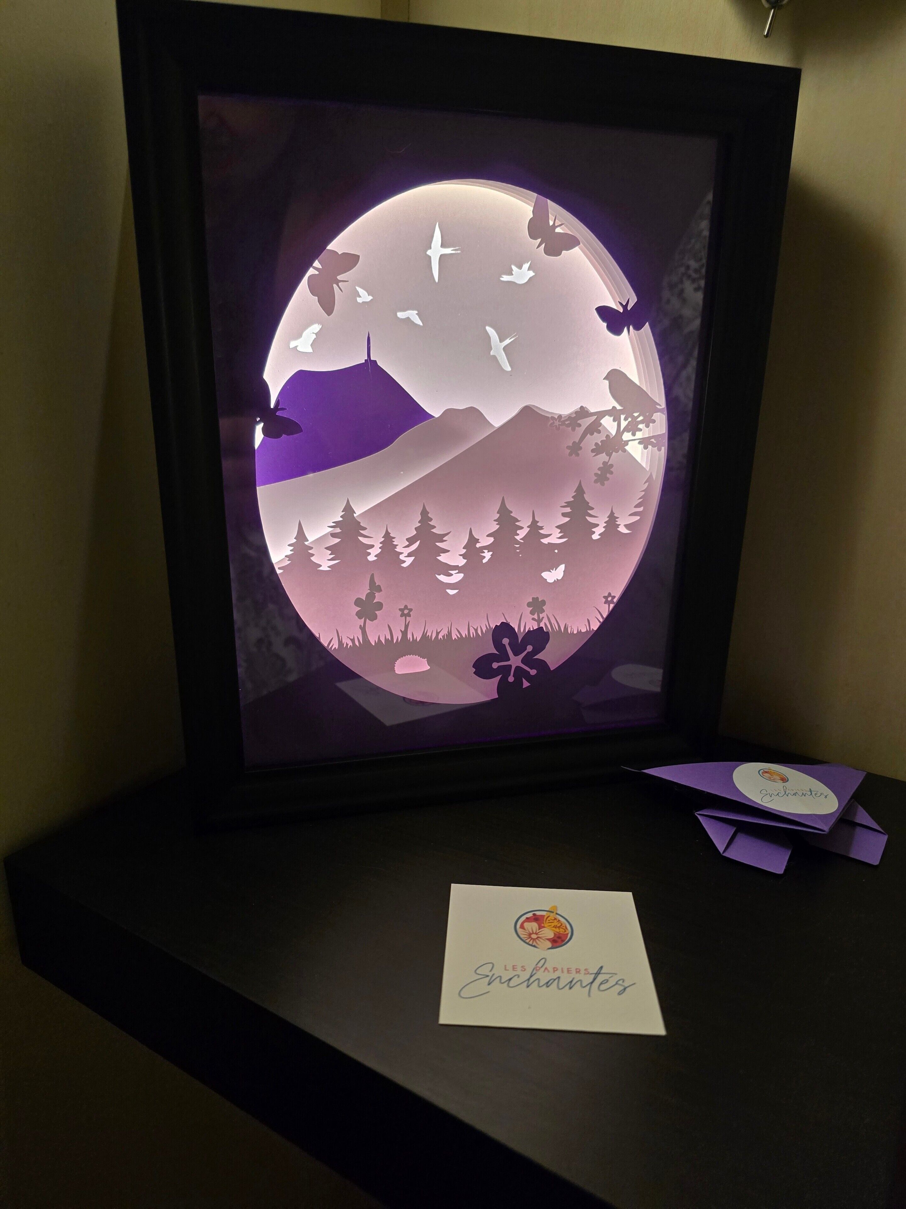 A framed artwork with a circular design featuring mountains, trees, and flying birds.