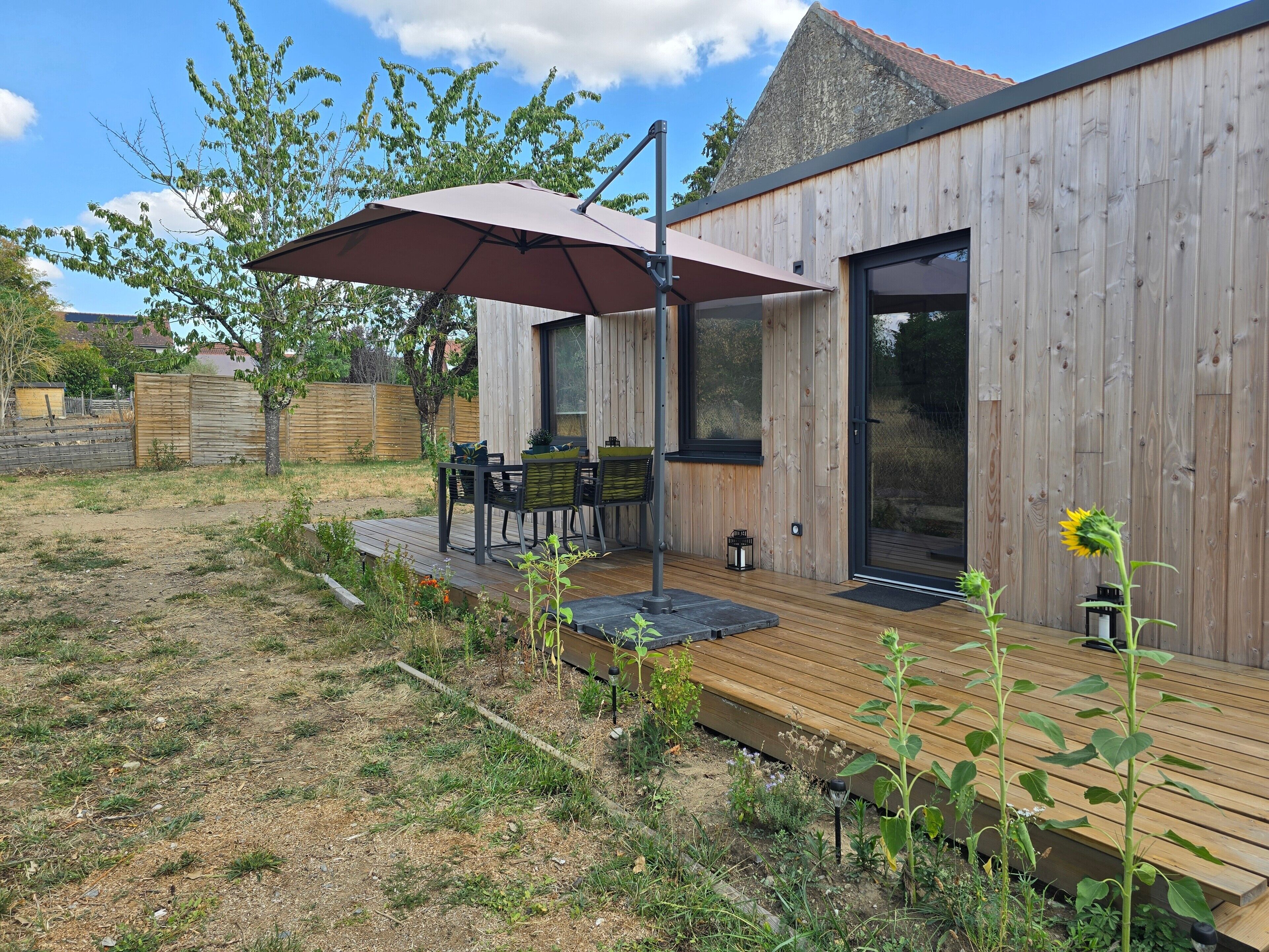 Une terrasse en bois avec une table et des chaises, un grand parasol et un jardin avec des plantes.