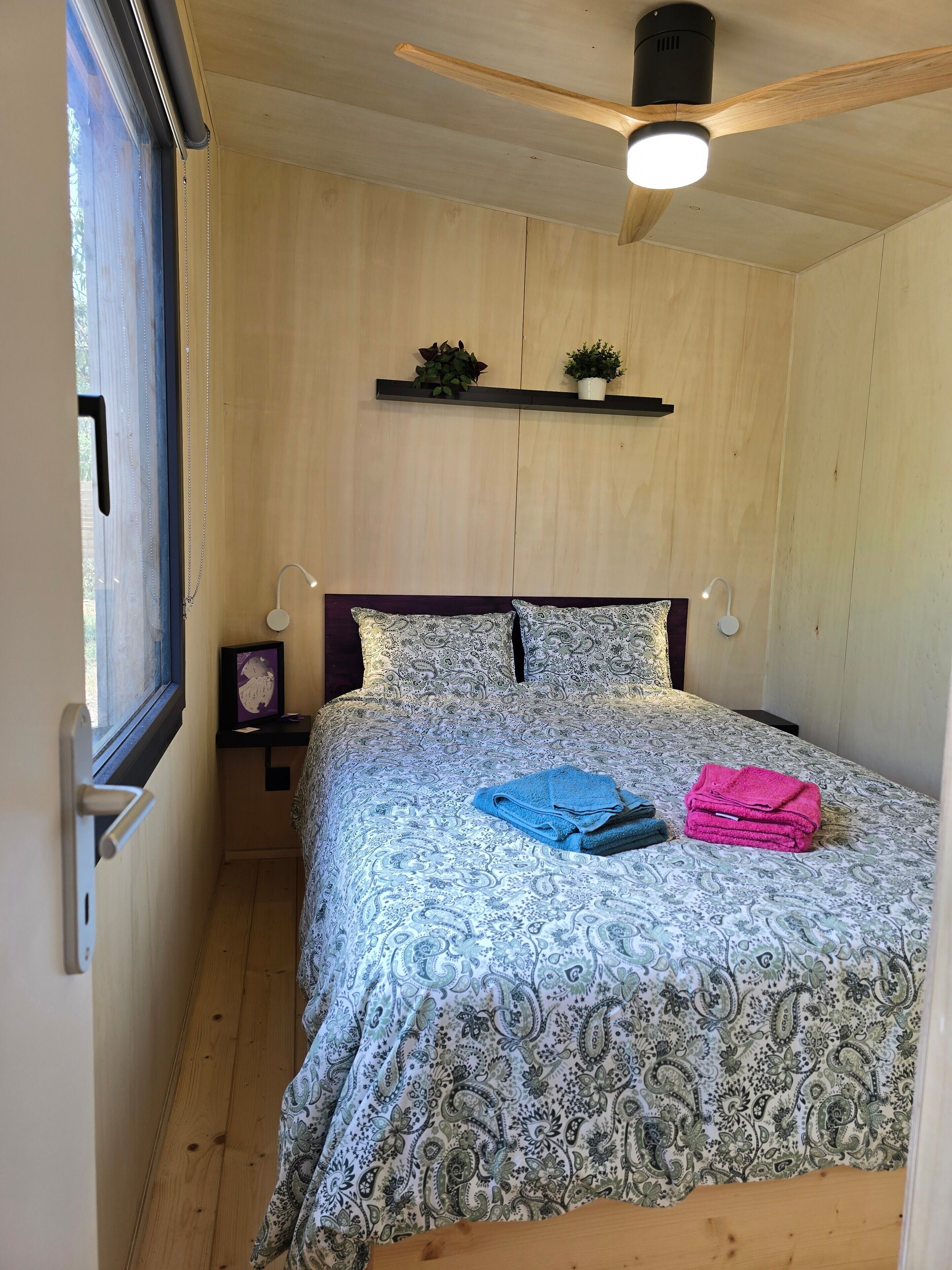A bedroom with a wooden ceiling fan, a bed with patterned bedding, a nightstand with a lamp, and a window with curtains.